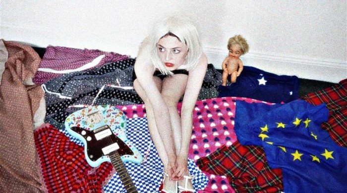 Girl in centre of image with blonde hair sitting on top of different blankets with a bass guitar to her left and a toy baby doll to her right.